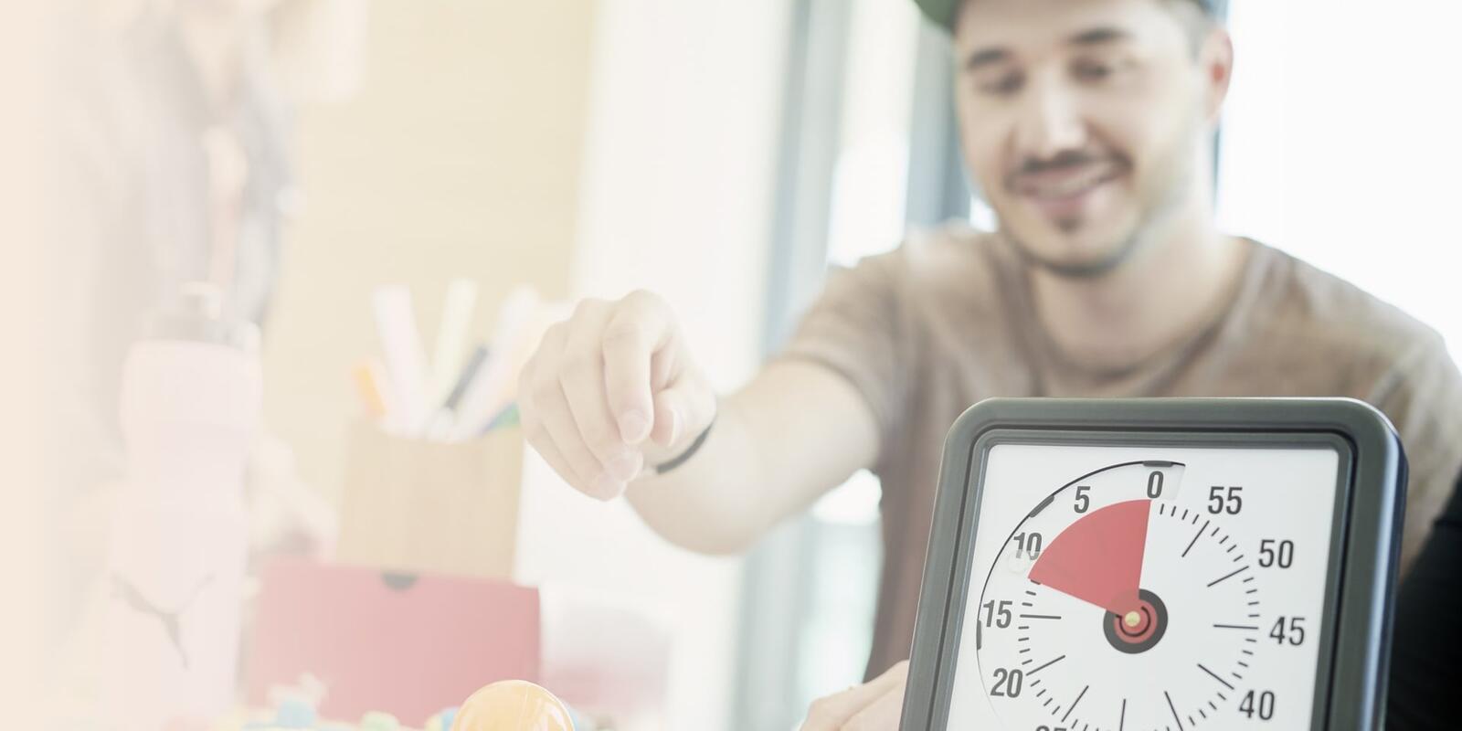 Eine Stoppuhr steht auf einem Tisch vor einem jungen Mann, der gerade nach etwas greift.